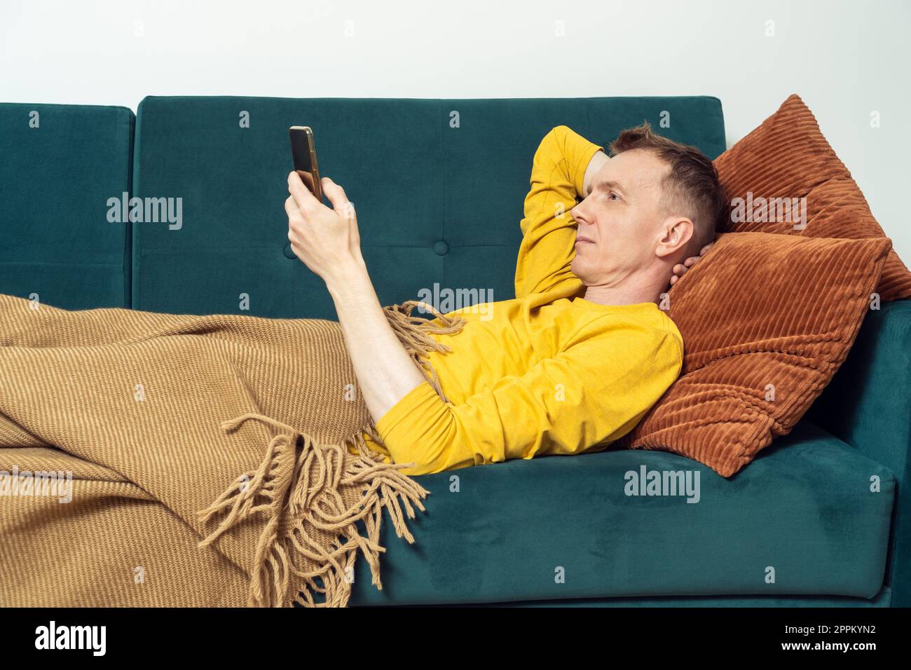 Ein erwachsener Mann liegt auf dem Sofa und chattet mit dem Smartphone, Seitenansicht. Person, die sich auf dem Kissen unter der Decke ruht und sich entspannen kann. Stockfoto