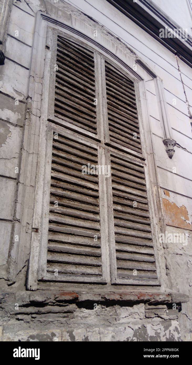 Geschlossene Fensterläden an einem hohen Fenster mit Plattbändern. Die Fassade des Gebäudes, die lange Zeit nicht repariert wurde. Der Putz blättert unter dem Fenster ab. Die Architektur der alten europäischen Stadt Stockfoto