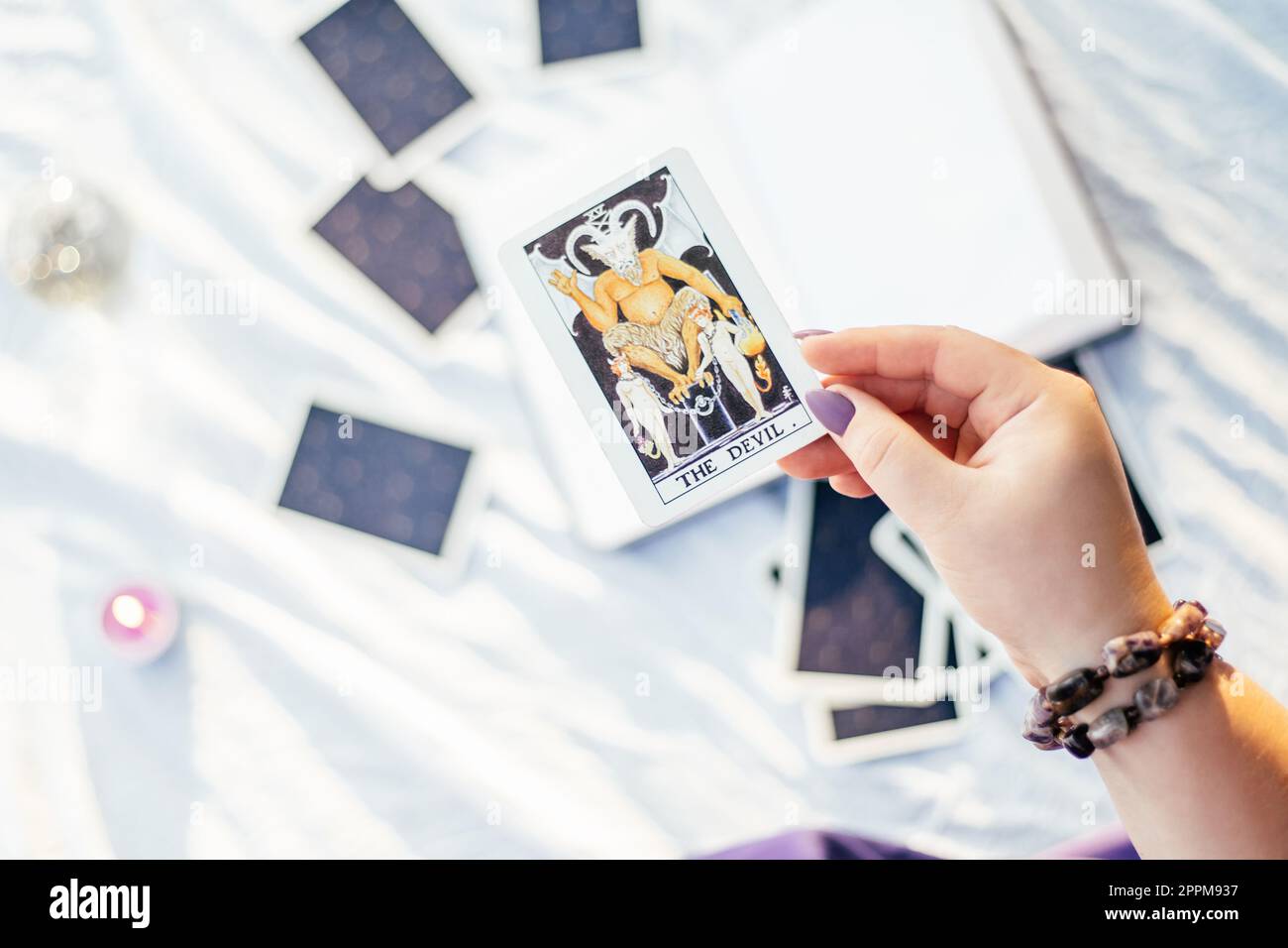 Eine weibliche Hand mit lila Nägeln hält die Tarotkarte mit dem Namen „Devil“ auf einer weißen Oberfläche mit offenem Notizbuch und Kerze. Draufsicht Stockfoto