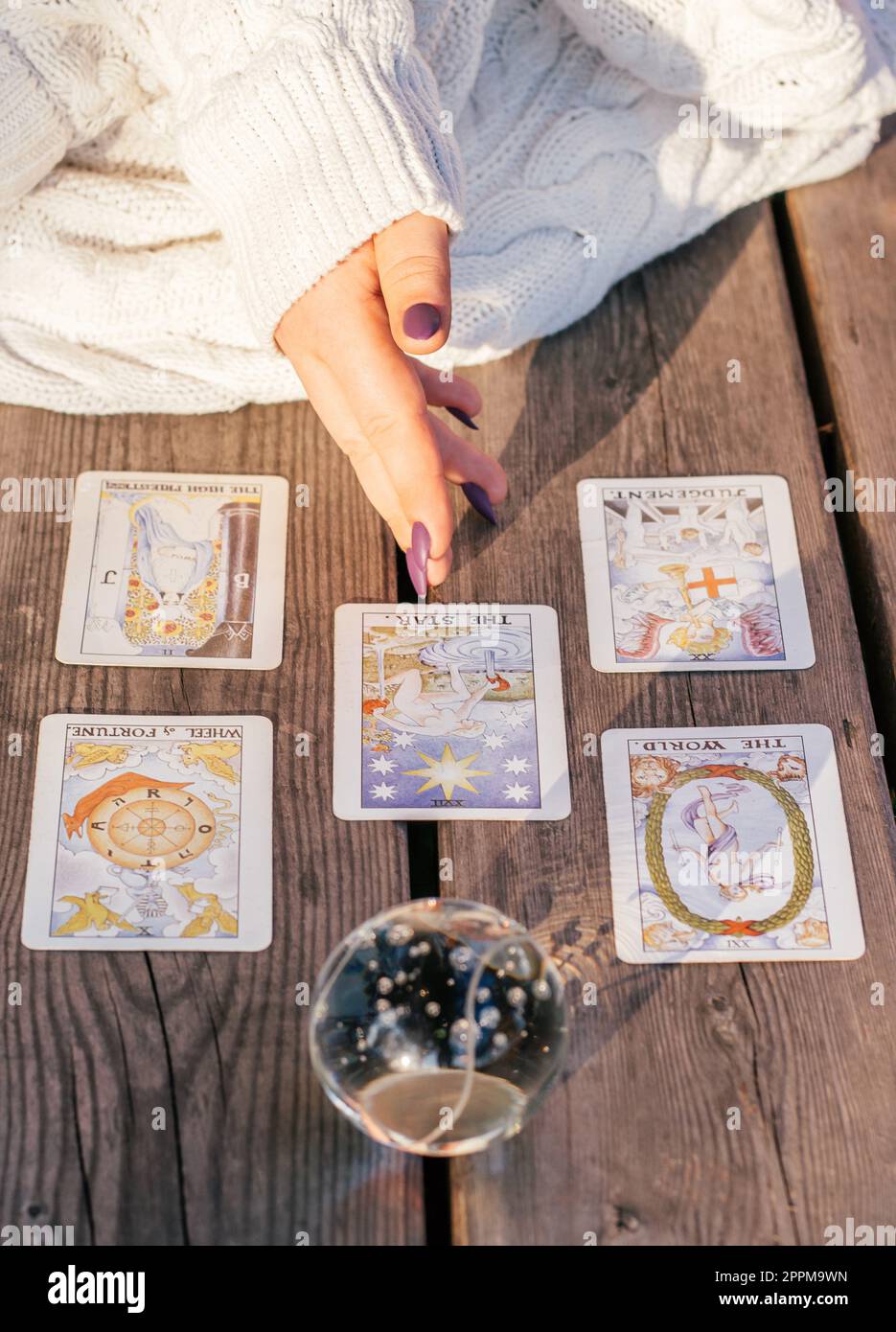 Die Hand einer Frau mit lila Nägeln zeigt auf fünf Tarot-Karten, die auf einer Holzfläche neben einer Kristallkugel verteilt sind. Vertikal Stockfoto