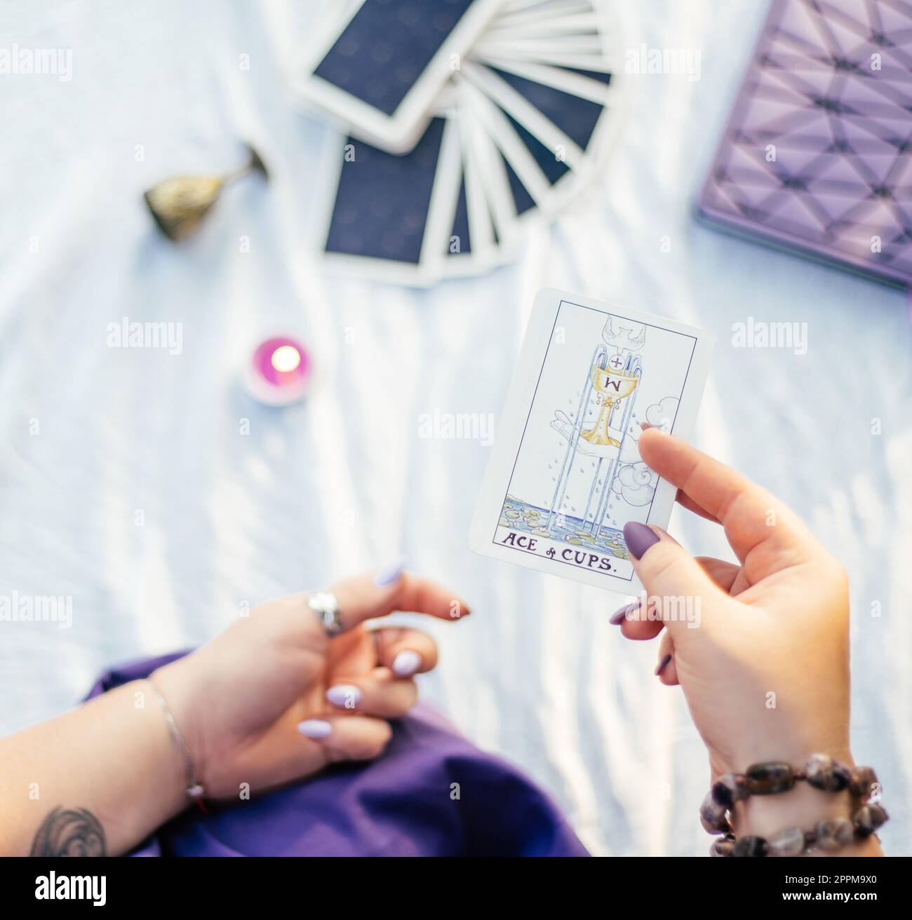 Eine weibliche Hand mit lila Nägeln hält eine Tarotkarte mit dem Namen „Ace of Cups“ auf einer weißen Oberfläche mit Notizbuch und Kerze. Draufsicht. Stockfoto