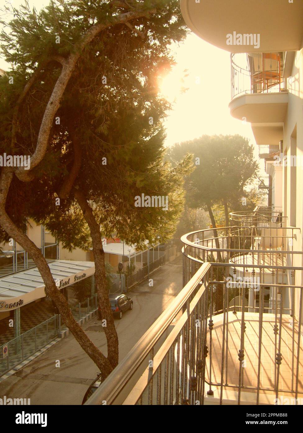 Rimini, Italien, 10. Dezember 2019 sonniger Wintermorgen auf dem Balkon eines Hotels in Italien. Mehrere Terrassen mit einem Metallzaun über einer schmalen italienischen Straße mit Kiefernbaum, gebadet in der gelben Sonne Stockfoto