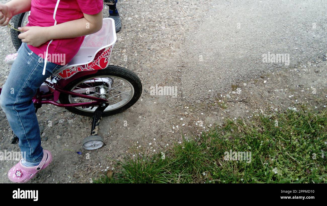 Belgrad, Serbien, 13. April 2020: Hinterrad eines Kinderfahrrads mit kleinem Zusatzrad. Teilweise sichtbar sind der Körper und das Bein eines Mädchens, das auf einem Fahrradsitz sitzt. Asphalt und grünes Gras Stockfoto