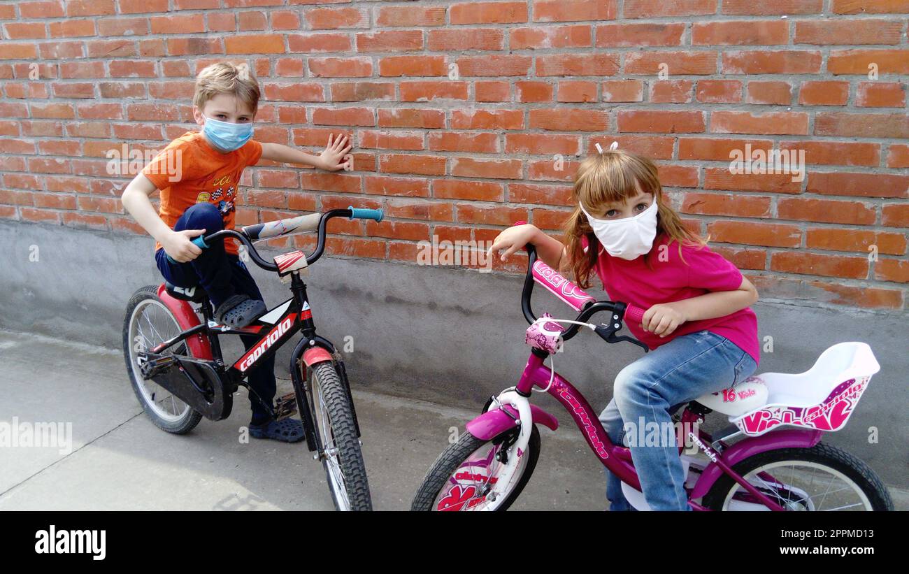 Kinder im Alter von 6 und 7 Jahren in weißen chirurgischen Schutzmasken gehen zum Radfahren. Pause beim Radfahren. Ein Mädchen in einem rosa T-Shirt und ein blonder Junge in Orange. Kinder auf der Straße, in der Nähe des alten Ziegelzauns. Stockfoto