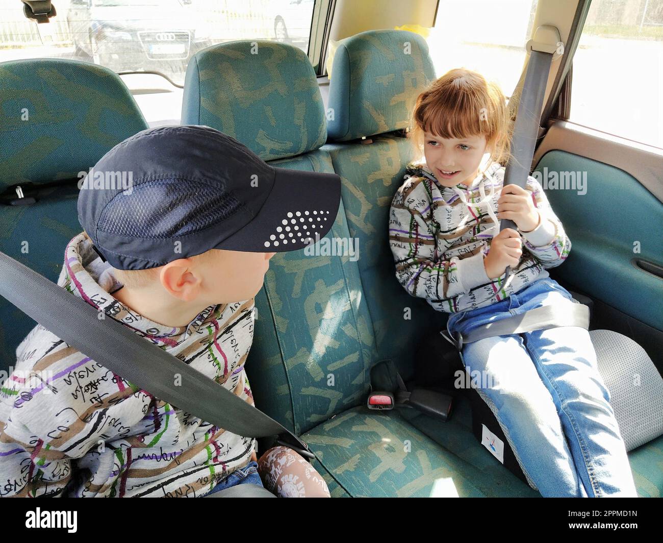 Kinder im Auto. Ein Junge und ein Mädchen sitzen auf dem Rücksitz, lachen und grimpern. Kinder schnallen sich an. Ein Kind mit Mütze. Zwillinge mit verschiedenen Gesichtern, die in derselben Kleidung gekleidet sind Stockfoto