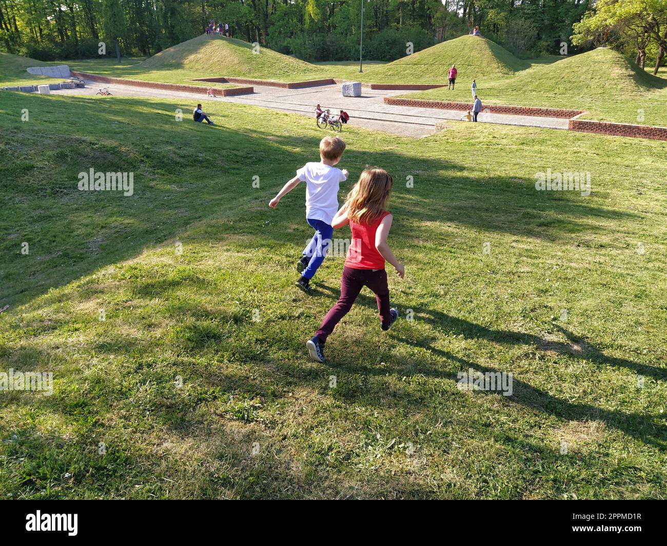 Sremska Mitrovica, Serbien - 24. April 2020 - Kinder Rennen im Park. Ein Junge in einem weißen T-Shirt und einer blauen Hose. Mädchen in einem roten Tanktop und einer Hose. Grüner Rasen oder Feld. Menschen gehen in einem Stadtpark Stockfoto