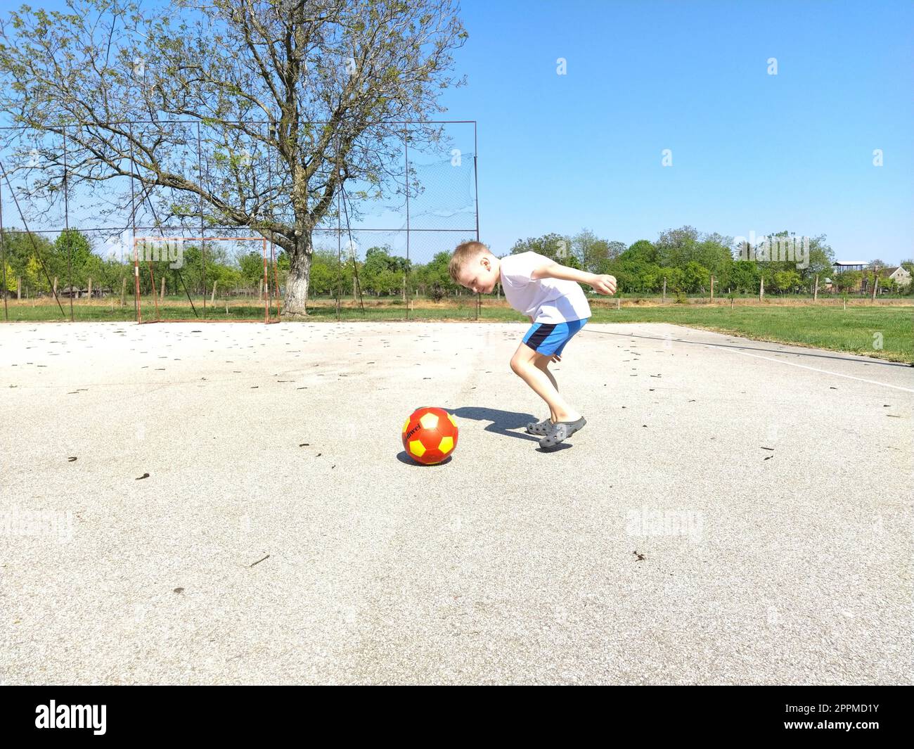 Sremska Mitrovica, Serbien. 6. Juni 2020. Ein Junge spielt Ball auf dem Spielplatz. Asphaltsportplatz. Ein Kind in einem weißen T-Shirt. Kleinkind mit blondem Haar, 7 Jahre alt. Laufen, Treten und Übungen Stockfoto