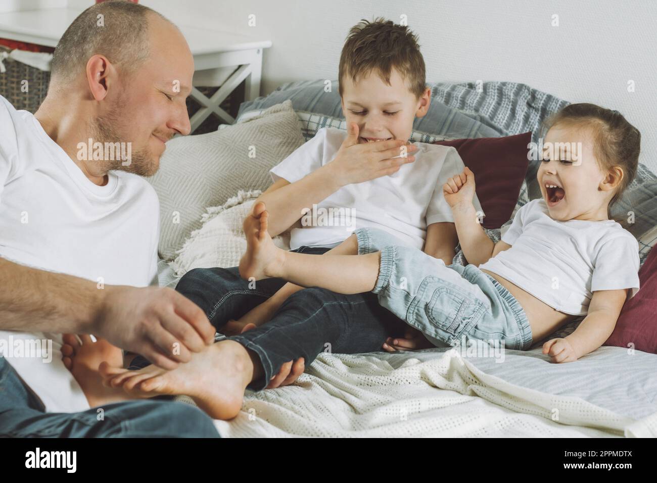 Vater spielt mit zwei Kindern im Bett. Dad kitzelt Kinderfüße. Familie von Daddy, Mädchen und Jungen Stockfoto
