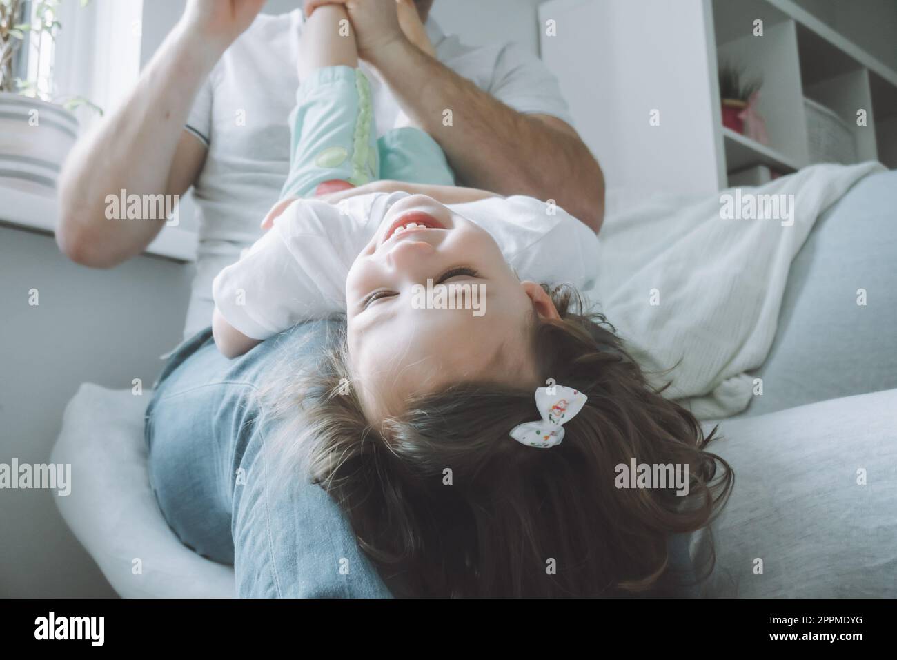Dad und kleines Mädchen spielen 2-4 auf der Couch. Das Kind liegt mit dem Kopf nach unten auf Vaters Schoß und lacht Stockfoto