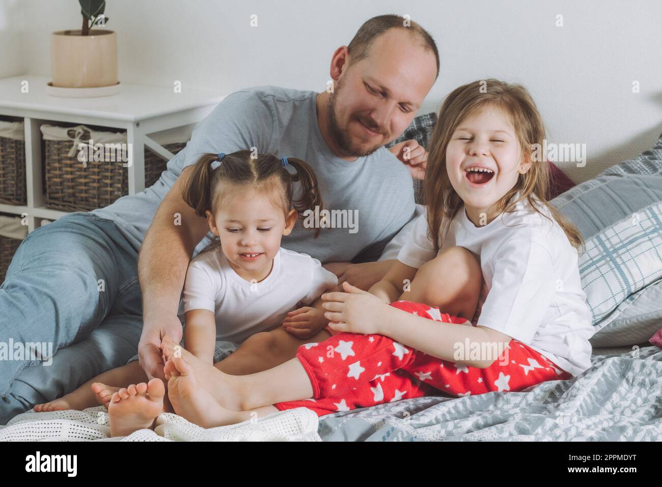 Vater spielt mit zwei Kindern im Bett. Dad kitzelt Kinderfüße. Eine Familie von Daddy und zwei kleine Mädchen Stockfoto