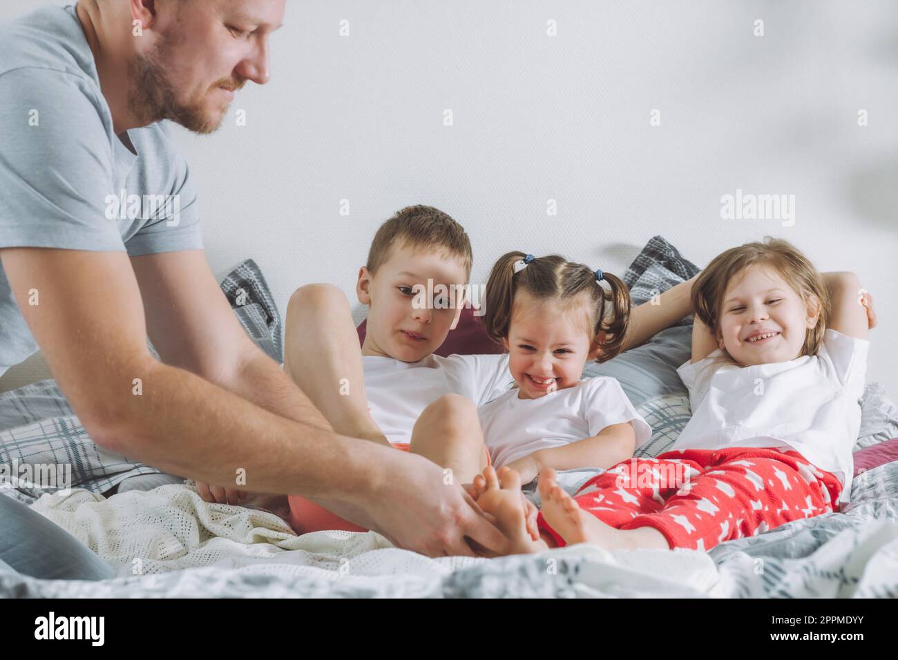 Vater spielt mit drei Kindern im Bett. Dad kitzelt Kinderfüße. Eine Familie von Daddy, zwei Mädchen und ein Junge Stockfoto