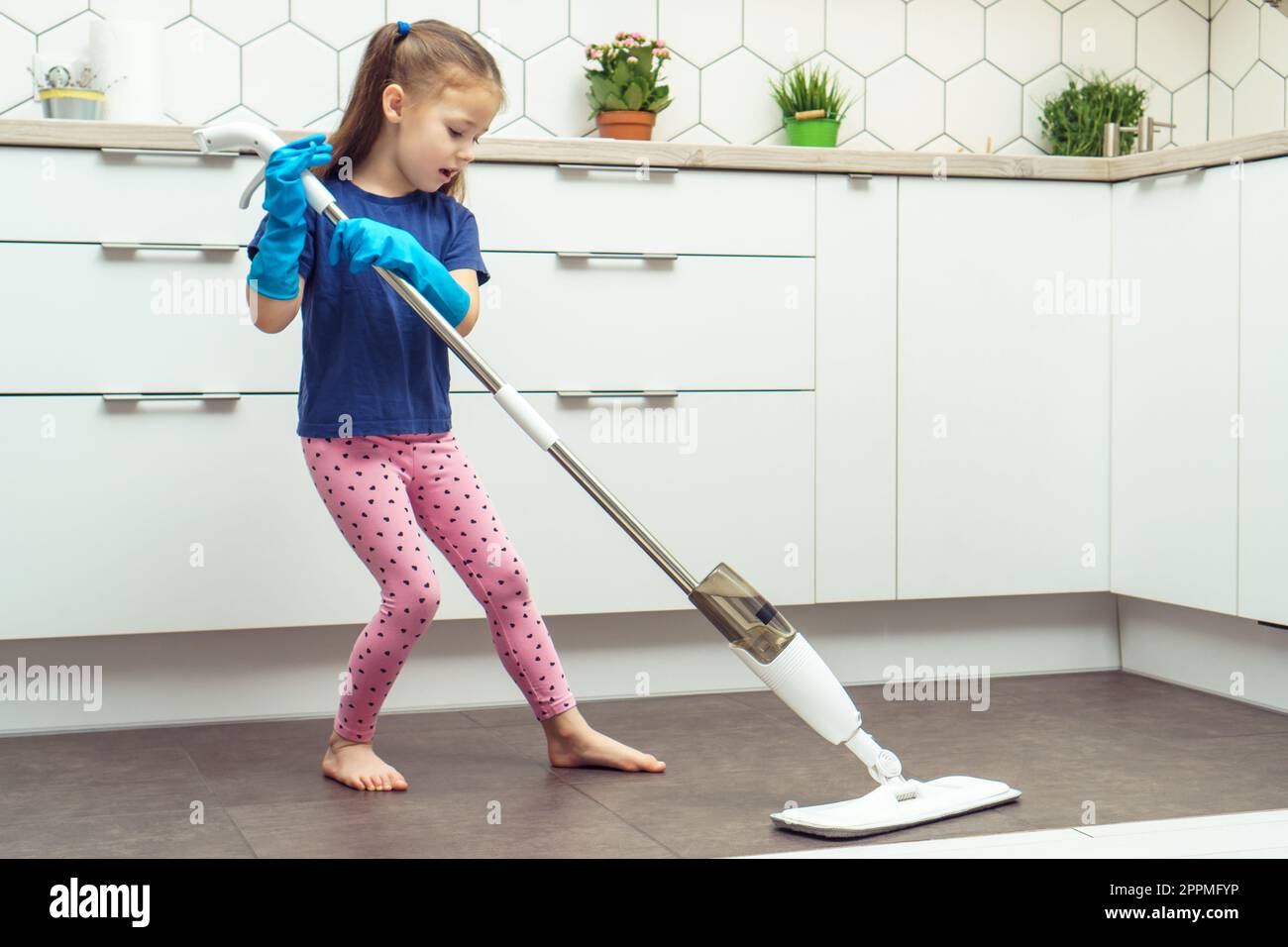 Ein hübsches kleines Mädchen in Haushaltshandschuhen wäscht den Boden mit einem modernen Mopp mit abnehmbarer Bürste in der Küche. Nassreinigung zu Hause. Stockfoto