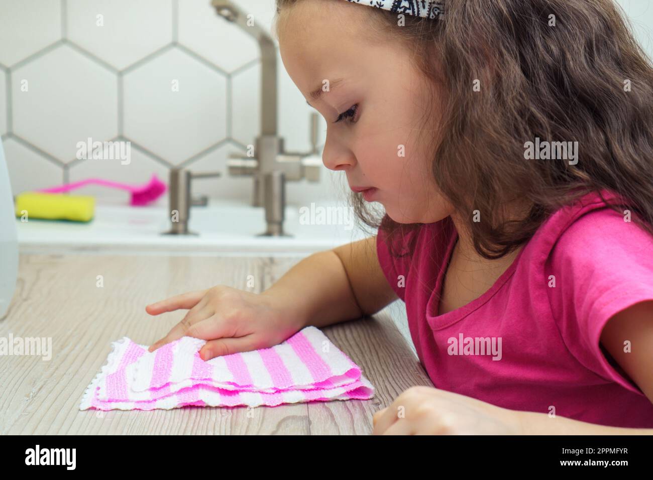 Hübsches kleines Mädchen, das den Küchentisch abwischte, mit einem Haushaltstuch zum Reinigen. Porträtaufnahme eines Kindes, das die Küche aufräumt. Stockfoto