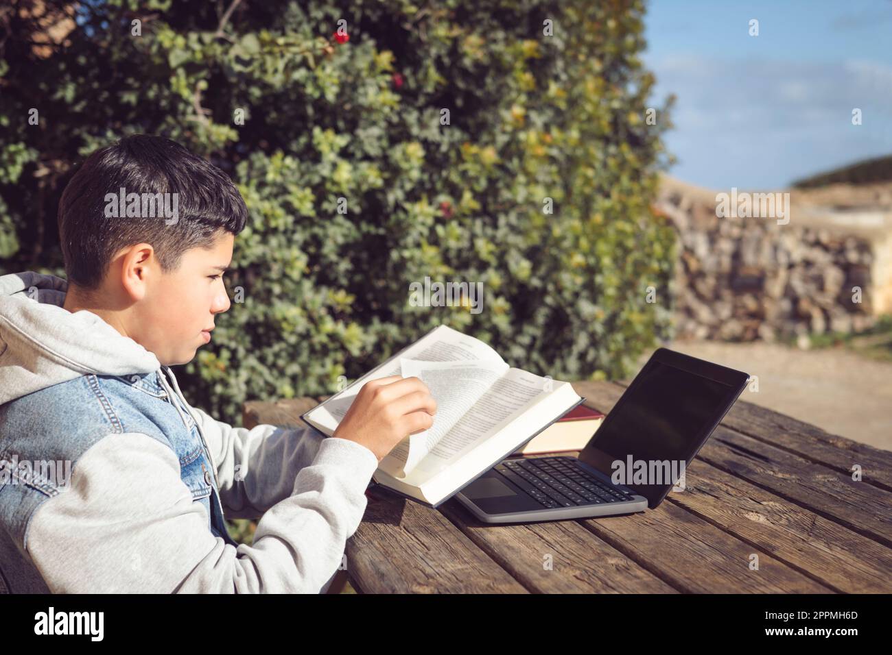 Junger Latinstudent blättert die Seite eines Buches vor einem Laptop um, draußen Stockfoto