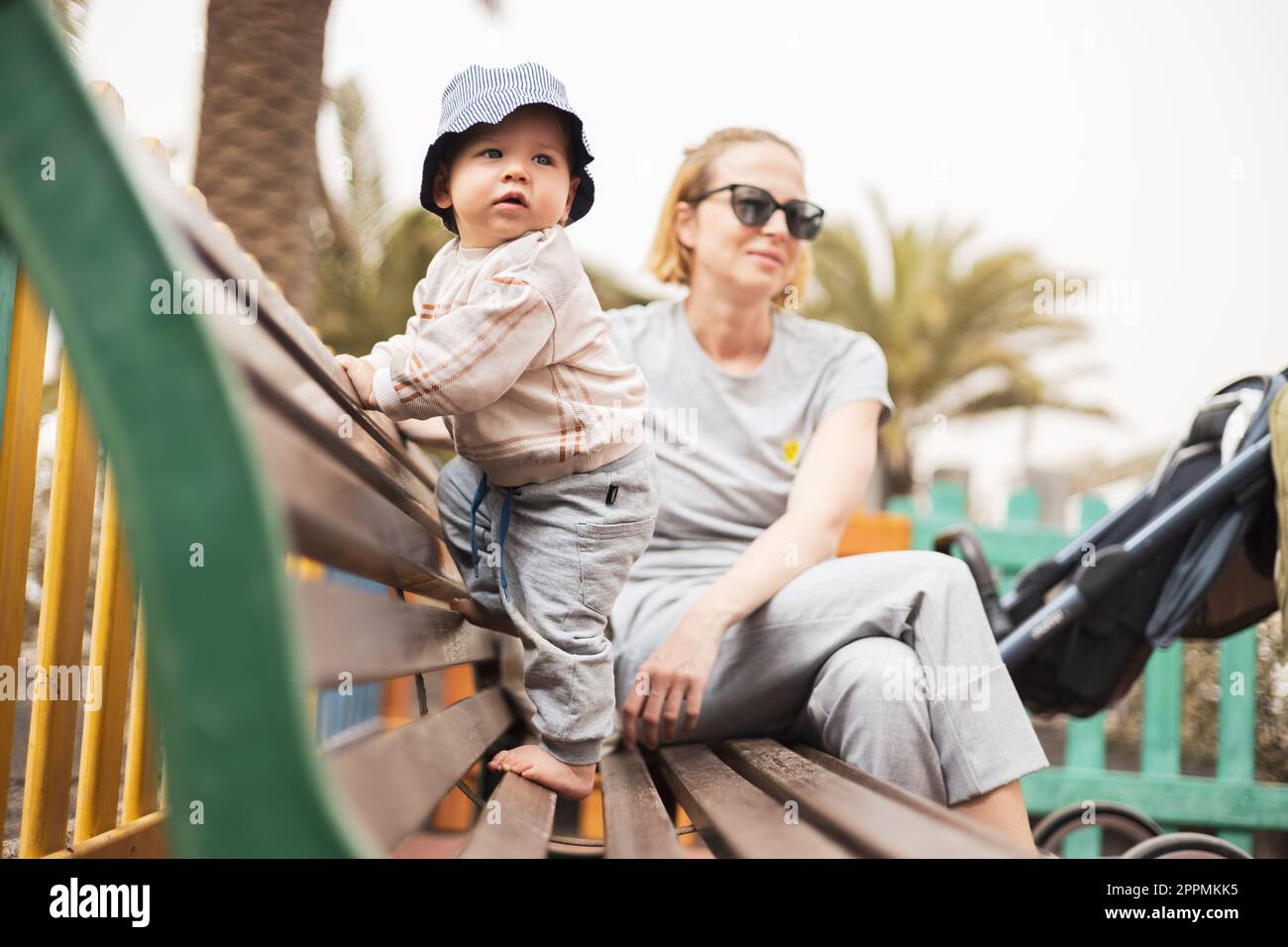 Junge Mutter mit ihrem süßen kleinen Jungen auf einer Bank auf dem Kinderspielplatz am warmen Sommertag. Stockfoto