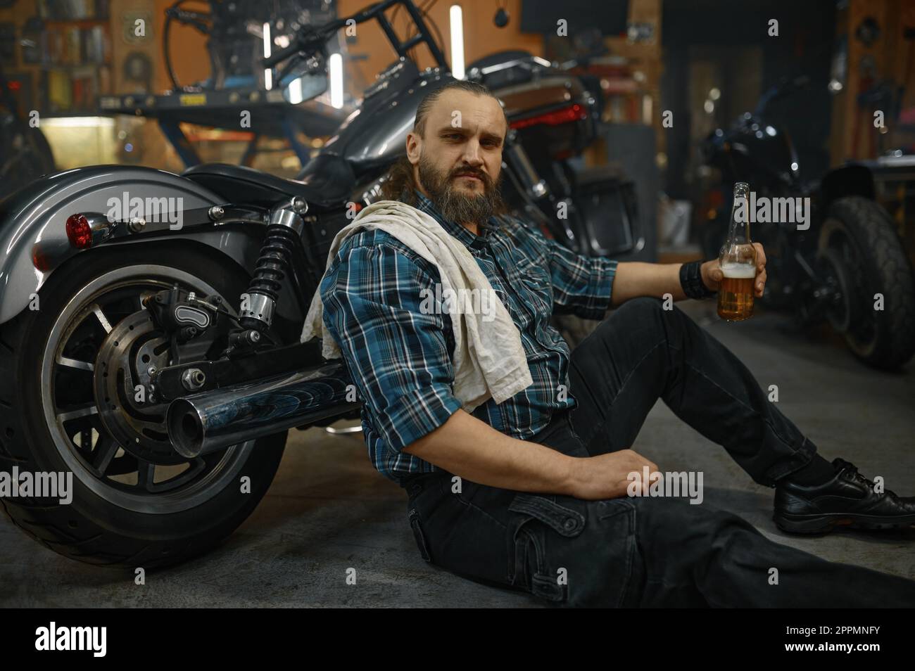 Erschöpfter, reifer Biker, der auf dem Boden sitzt und ein eröffnetes Glas Bier hält Stockfoto