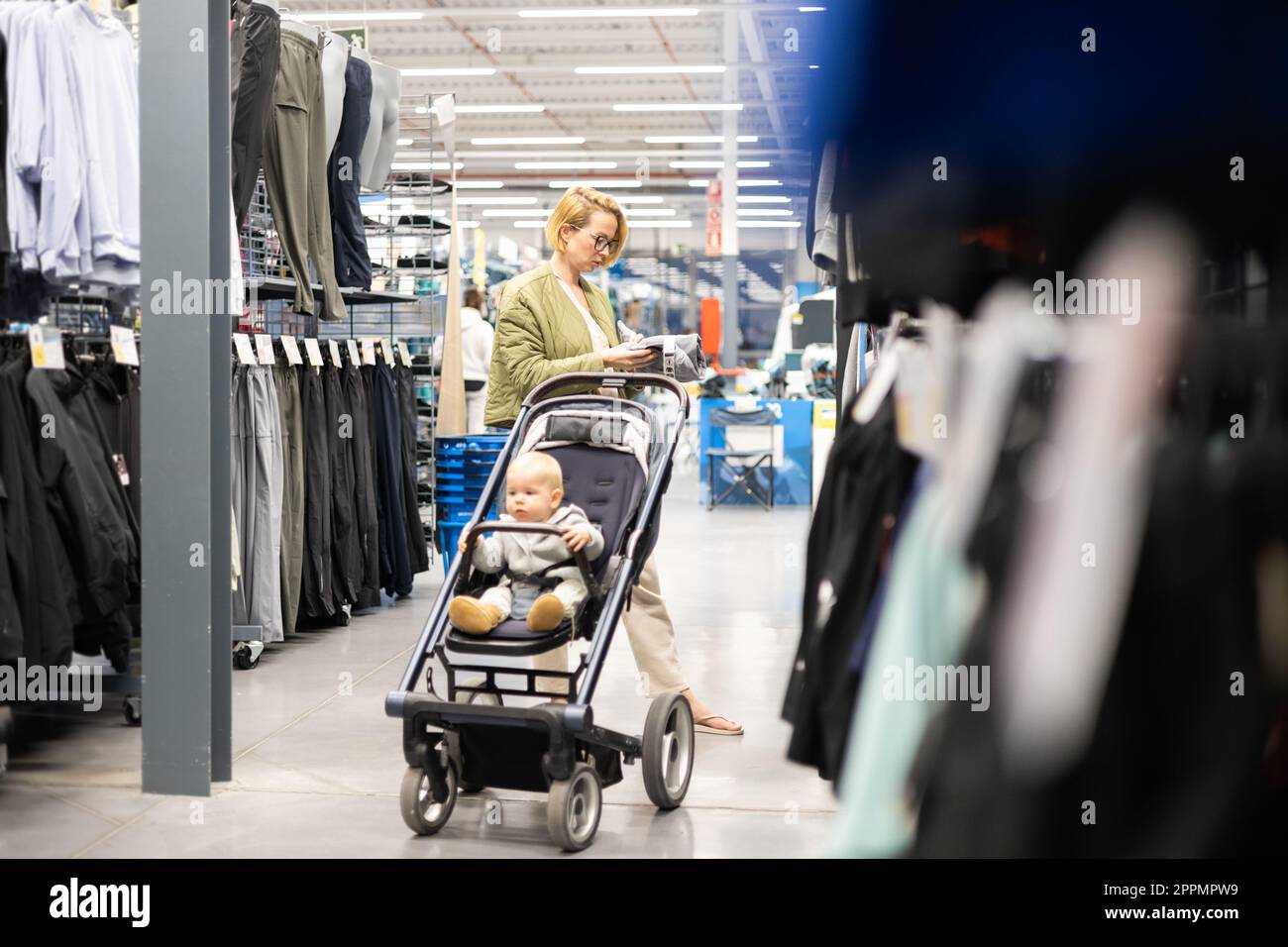 Lässig gekleidete Mutter, die in der Sportabteilung des Supermarkts Sportschuhe und Bekleidungsartikel aussucht, mit ihrem kleinen Jungen im Kinderwagen. Stockfoto