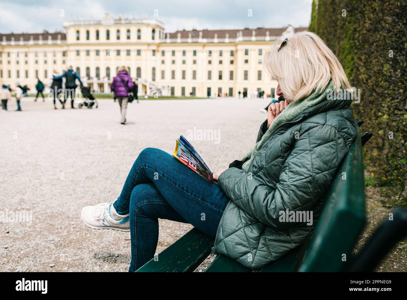 Blonde Frau in warmer Jacke, die im Frühling auf einer Bank im Park des Schlosses Schönbrunn sitzt und einen Reiseführer, Wien, Österreich, liest Stockfoto