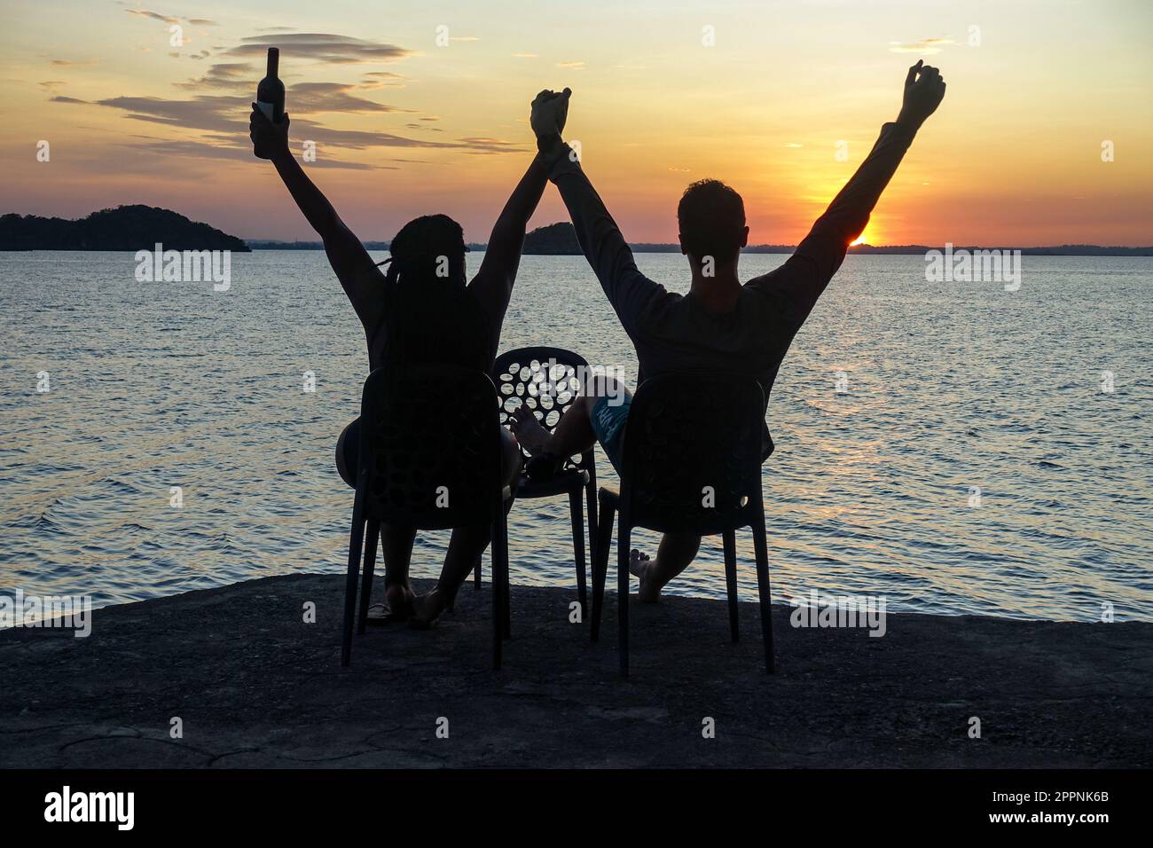 Ein Paar beobachtet den Sonnenuntergang auf den Philippinen, sitzt auf Stühlen und hebt die Arme mit einer Flasche Wein. Aufgenommen auf den 100 Inseln. Stockfoto