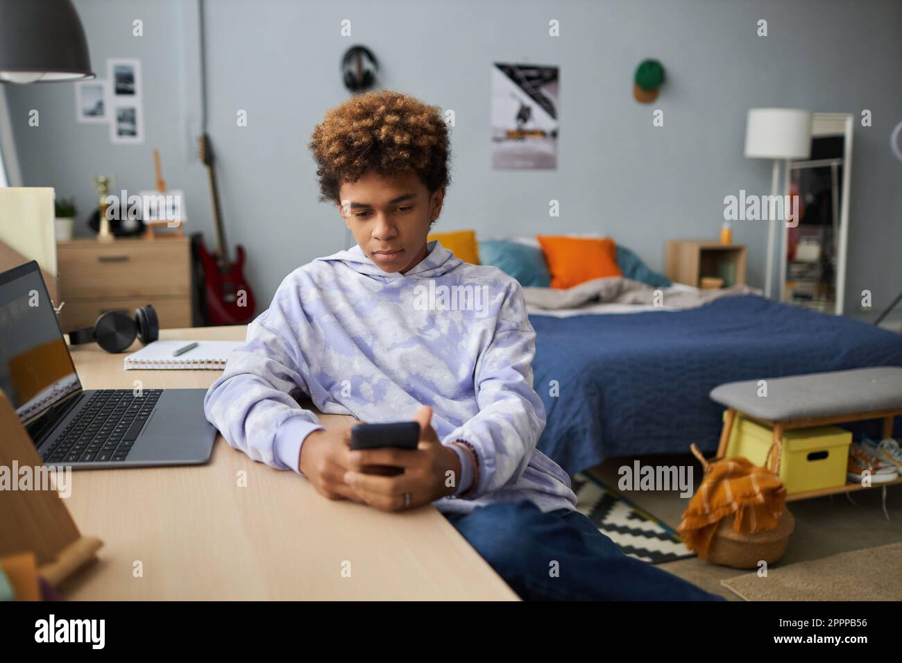 Ein echter Schuljunge in einem Hoodie, der auf den Smartphone-Bildschirm schaut, während er sich mit dem Laptop ein Online-Video am Schreibtisch ansieht und sich dabei ein Doppelbett mit Kissen ansieht Stockfoto
