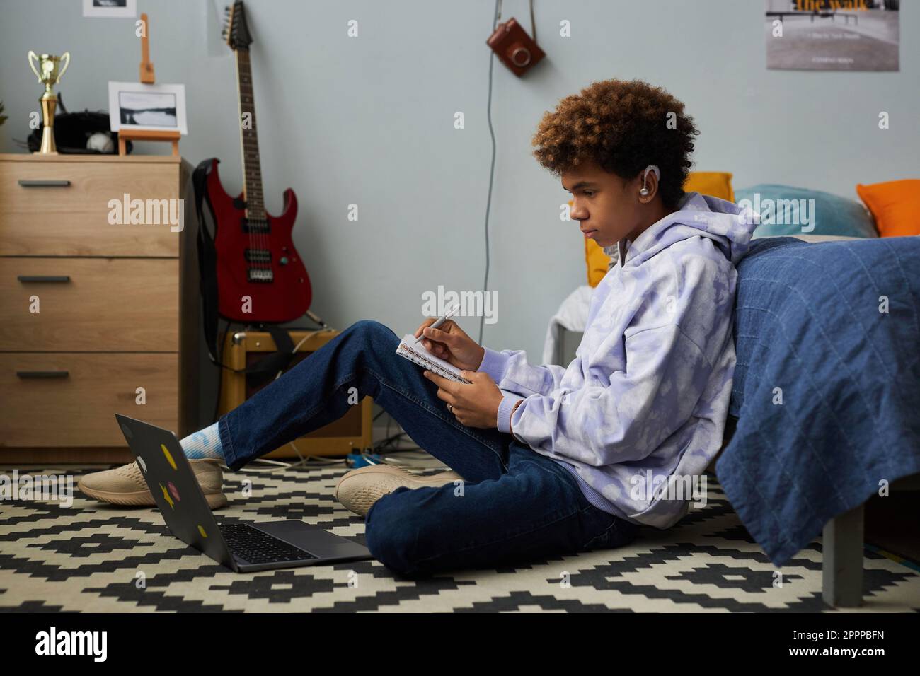 Afroamerikanischer Teenager mit Kopfhörern, der sich während einer Online-Vorlesung Notizen im Copybook macht, während er vor einem Laptop auf dem Boden neben seinem Bett sitzt Stockfoto