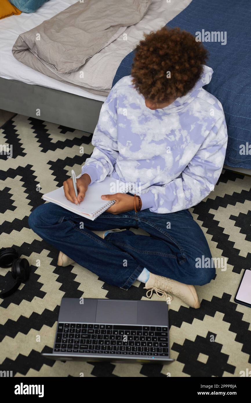 Der große Winkel eines afroamerikanischen Teenager-Schuljungen, der sich Notizen in einem Copybook macht, während er auf dem Boden sitzt und sich auf das Schulseminar vorbereitet Stockfoto