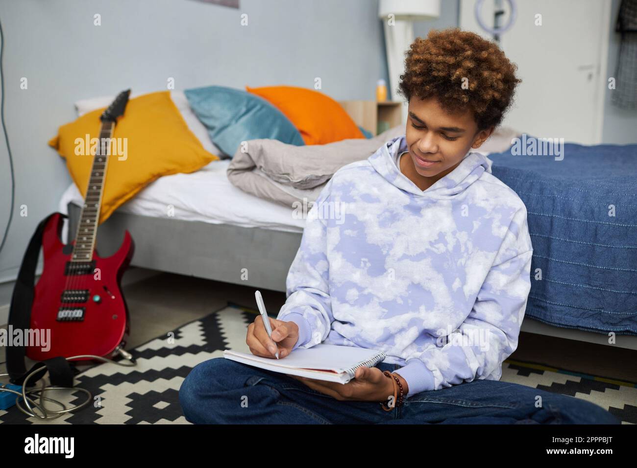 Jugendlicher Student in Freizeitkleidung, der sich Notizen in einem Copybook macht, während er auf dem Boden mit einem Doppelbett sitzt und die Hausaufgabe vorbereitet Stockfoto