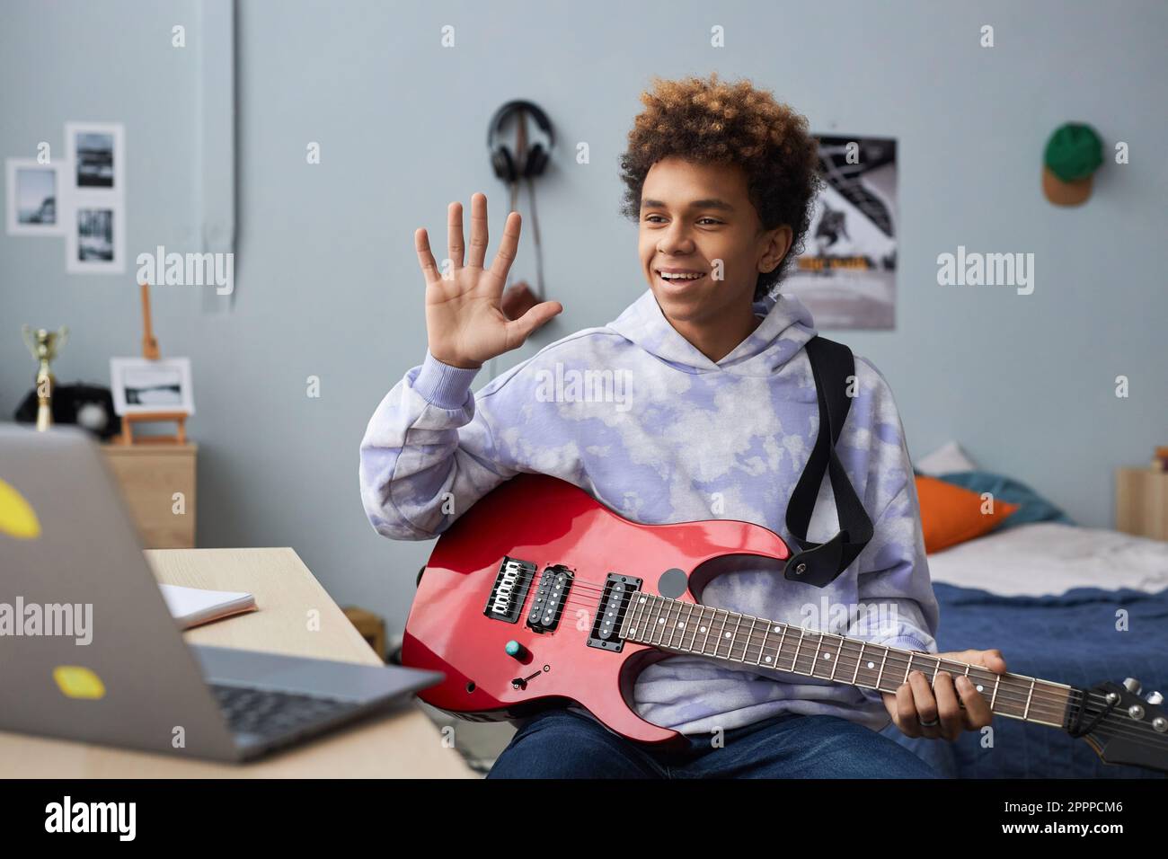 Glücklicher Teenager in Freizeitkleidung begrüßt seinen Musiklehrer, indem er vor dem Online-Unterricht mit der Hand wedelt, während er vor dem Laptop auf dem Bett sitzt Stockfoto