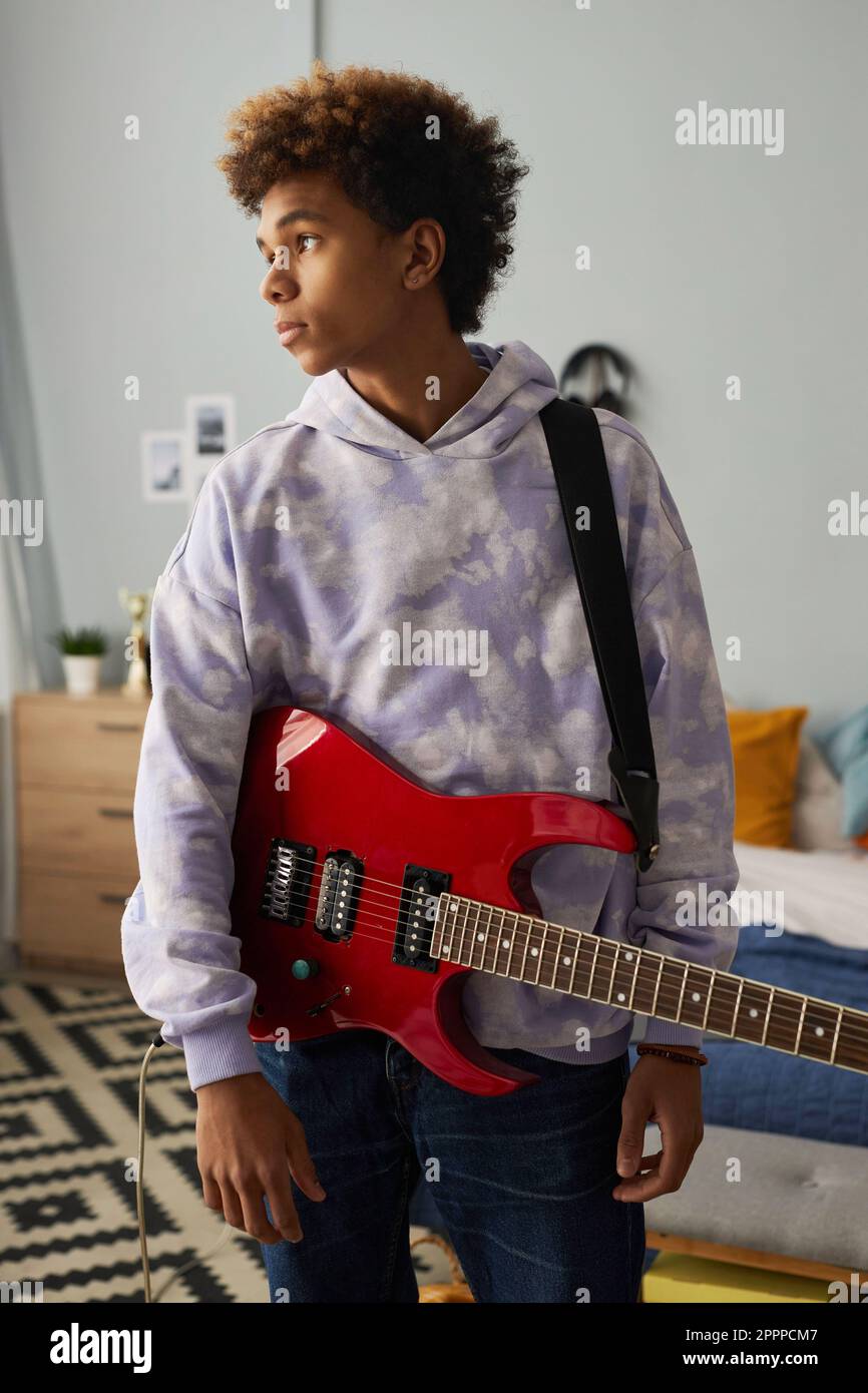 Ein heiterer Teenager mit einer elektrischen Gitarre, die an seiner Schulter hängt, vor der Kamera steht und in der Wohnung zur Seite schaut Stockfoto