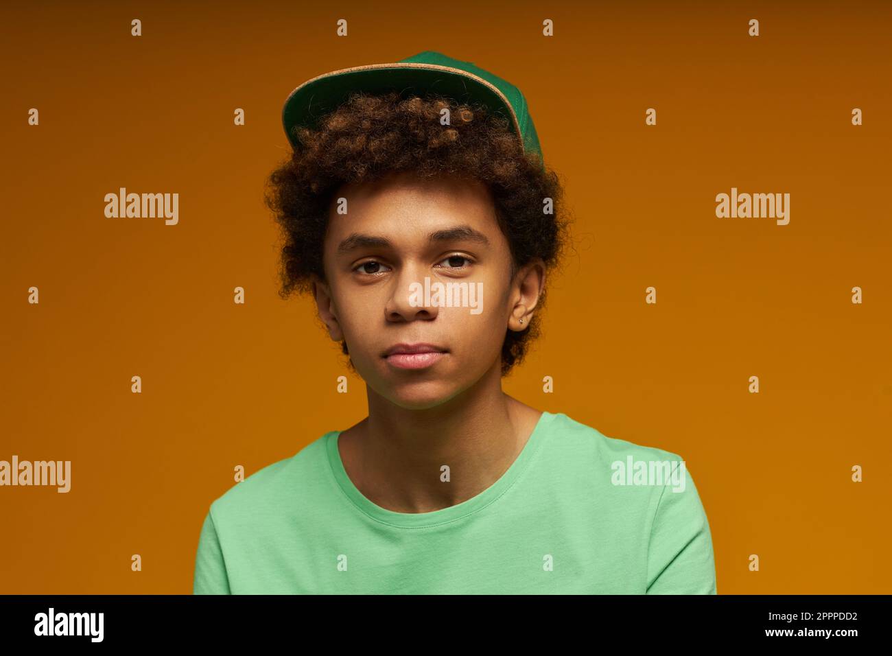 Süßer Teenager mit neutralem Gesichtsausdruck trägt ein grünes T-Shirt und eine Kappe und schaut in die Kamera, während er isoliert auf gelbem Hintergrund posiert Stockfoto