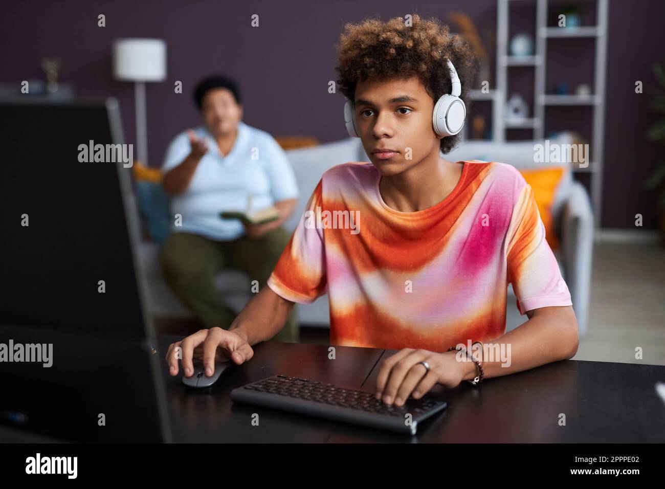 Ein gewissenhafter Teenager mit Kopfhörern klickt mit der Maus und tippt auf der Computertastatur, während er am Schreibtisch sitzt und Hausaufgaben macht Stockfoto