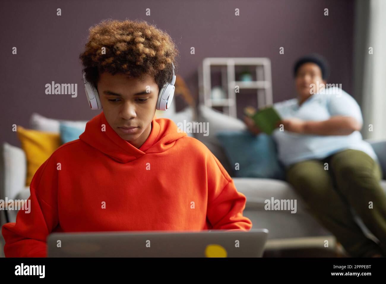 Konzentrieren Sie sich auf den fleißigen Schuljungen mit Kopfhörern und rotem Hoodie, der vor dem Laptop sitzt und Online-Videos ansieht oder Hausaufgaben vorbereitet Stockfoto