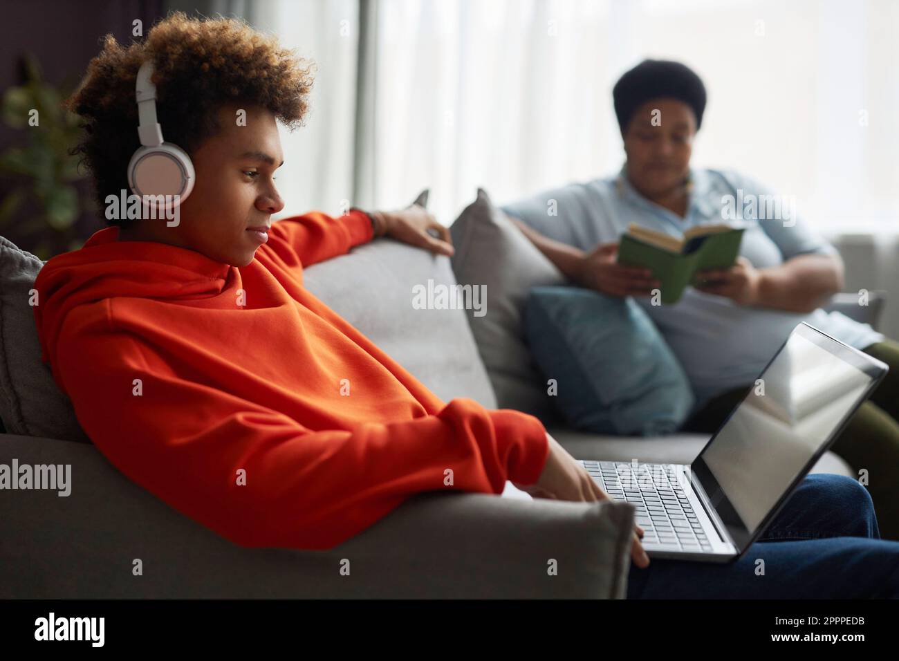 Seitenansicht eines Jungen im Teenageralter mit Kopfhörern und rotem Hoodie, der sich einen Online-Film oder ein Video auf dem Laptop-Bildschirm ansieht, während eine Frau ein Buch liest Stockfoto