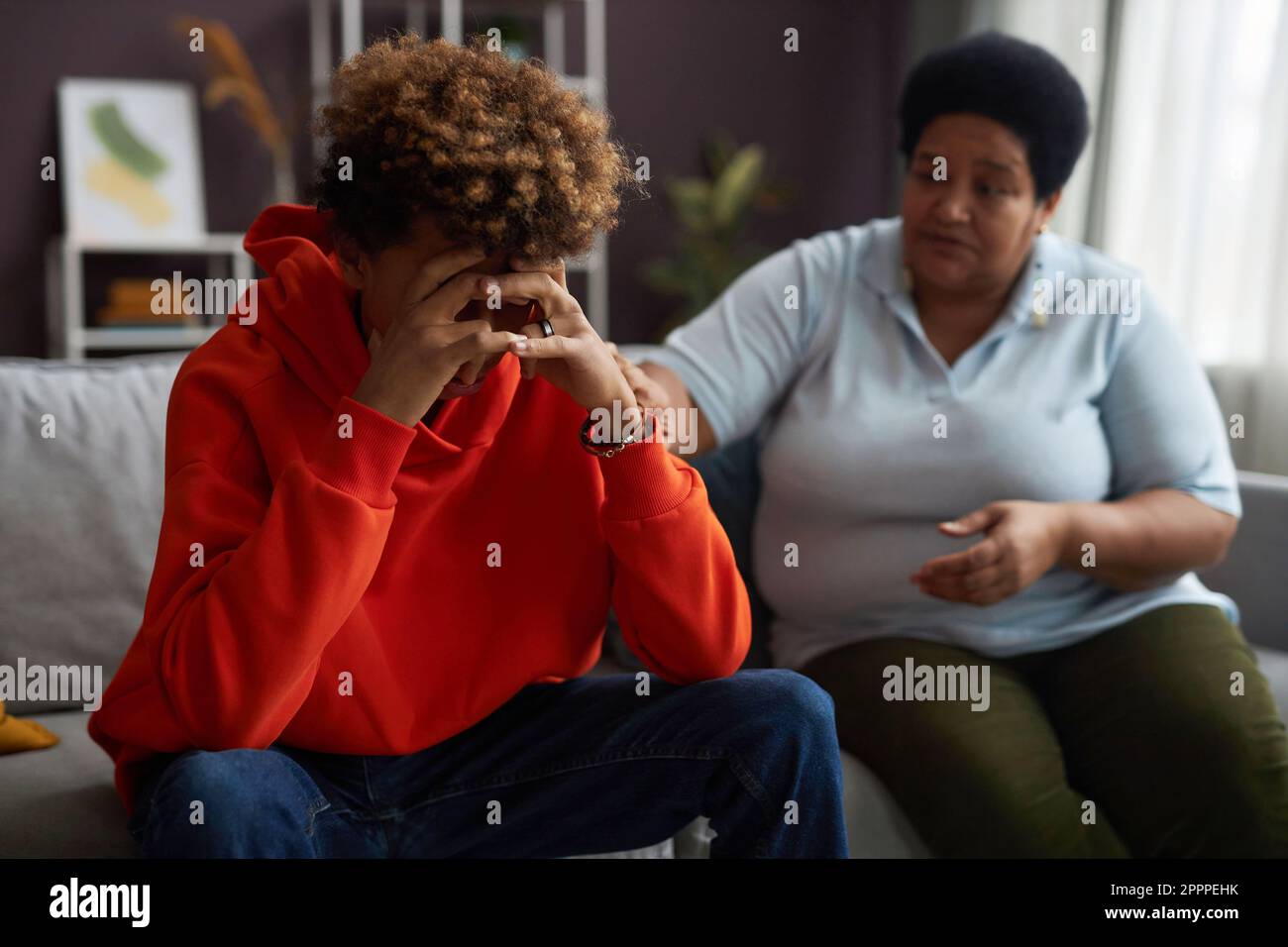 Konzentrieren Sie sich darauf, dass ein gestresster Teenager sein Gesicht in den Händen versteckt, während er vor der Kamera auf der Couch sitzt, während eine Erwachsene Frau ihn unterstützt Stockfoto