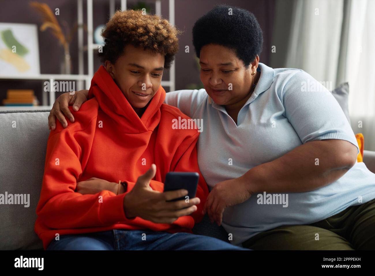 Ein afroamerikanischer Teenager in rotem Hoodie zeigt seiner Großmutter Online-Videos auf dem Smartphone, während beide auf dem Sofa sitzen Stockfoto