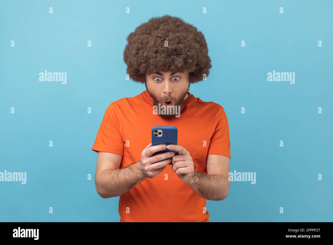 Porträt eines erstaunten Mannes mit Afro-Frisur, der ein orangefarbenes T-Shirt trägt, einen Post in einem sozialen Netzwerk mit Handy liest, und sich überrascht auswirkt. Innenstudio, isoliert auf blauem Hintergrund. Stockfoto