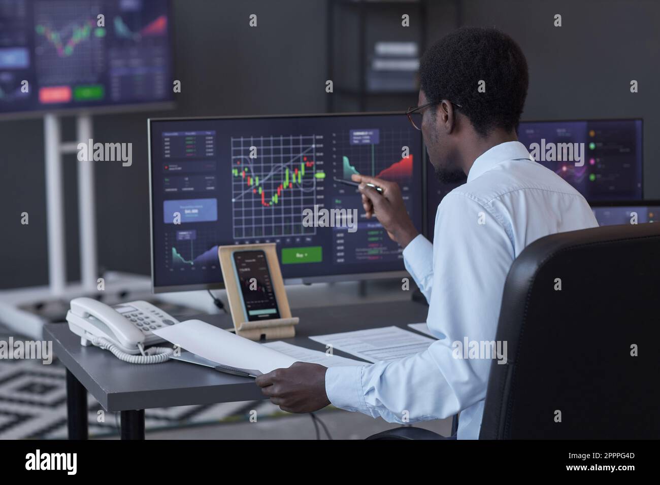 Rückansicht eines afroamerikanischen Brokers, der die Börse auf Computerbildschirmen analysiert, während er im Büro am Tisch sitzt Stockfoto