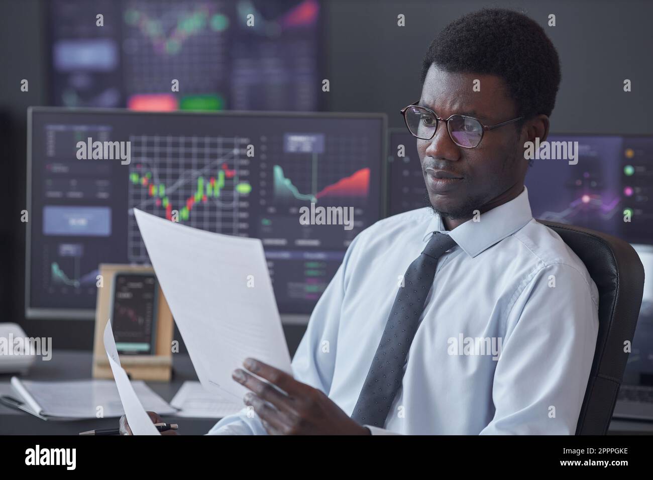 Ein afroamerikanischer Geschäftsmann mit Brille, der Finanzdokumente prüft, während er mit Computermonitoren mit Karten im Hintergrund am Tisch sitzt Stockfoto