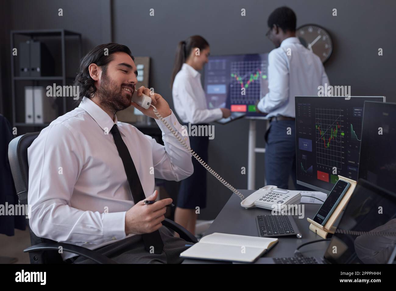 Junger Makler, der an seinem Arbeitsplatz vor Computermonitoren sitzt und am Telefon mit dem Kunden spricht Stockfoto