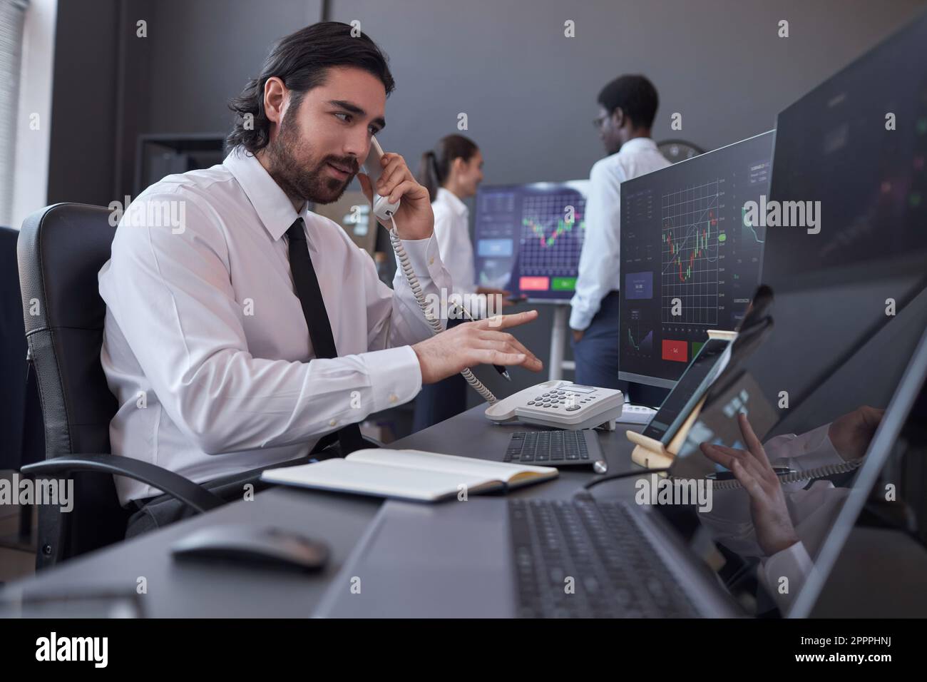 Ein junger Broker, der einen Kunden berät, indem er am Arbeitsplatz auf Computerbildschirme mit Karten zeigt Stockfoto