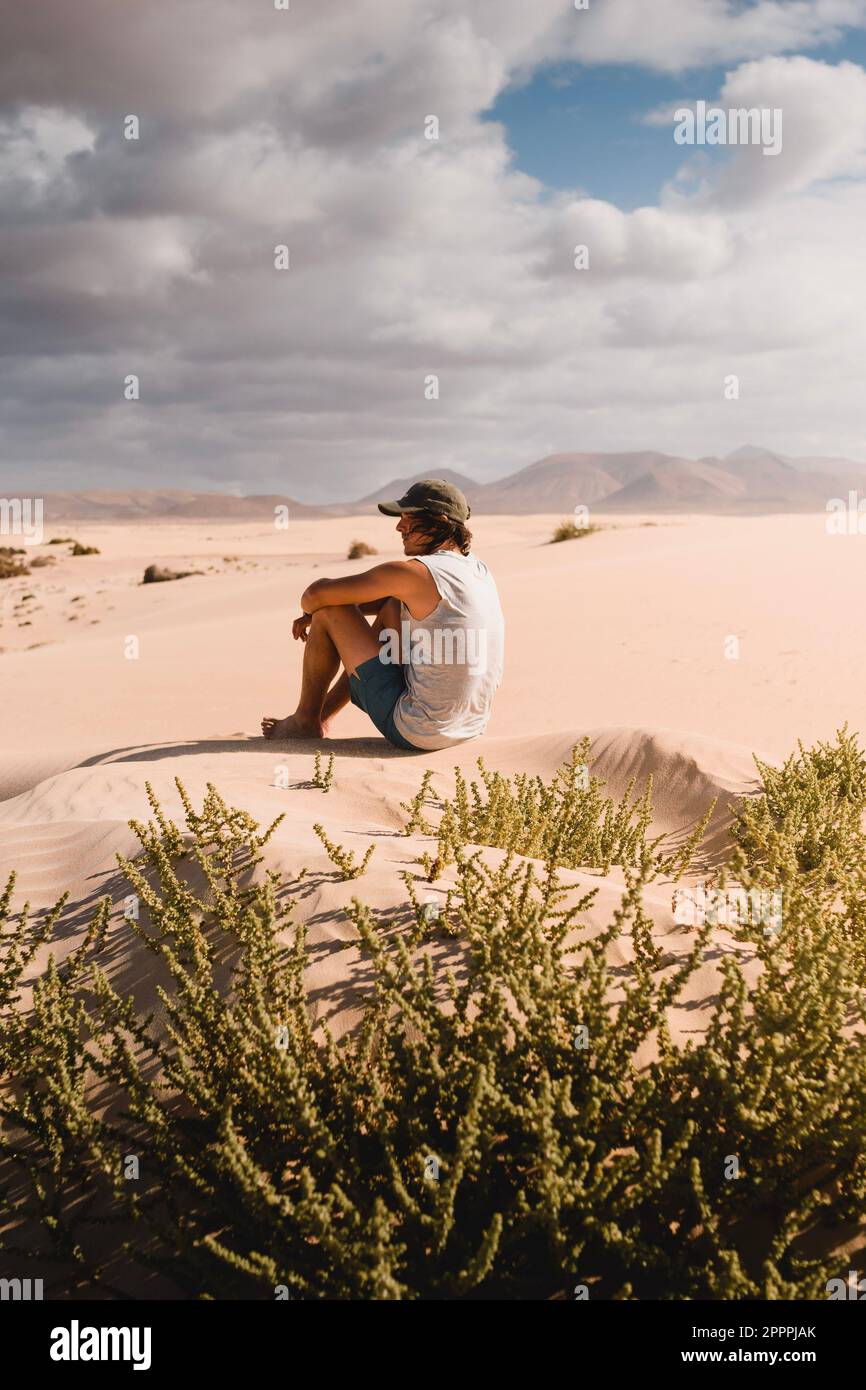 Junger Mann mit Mütze, der auf dem Wüstensand der Dünen von Corralejo in Fuerteventura sitzt und während einer Urlaubsreise den Horizont betrachtet Stockfoto