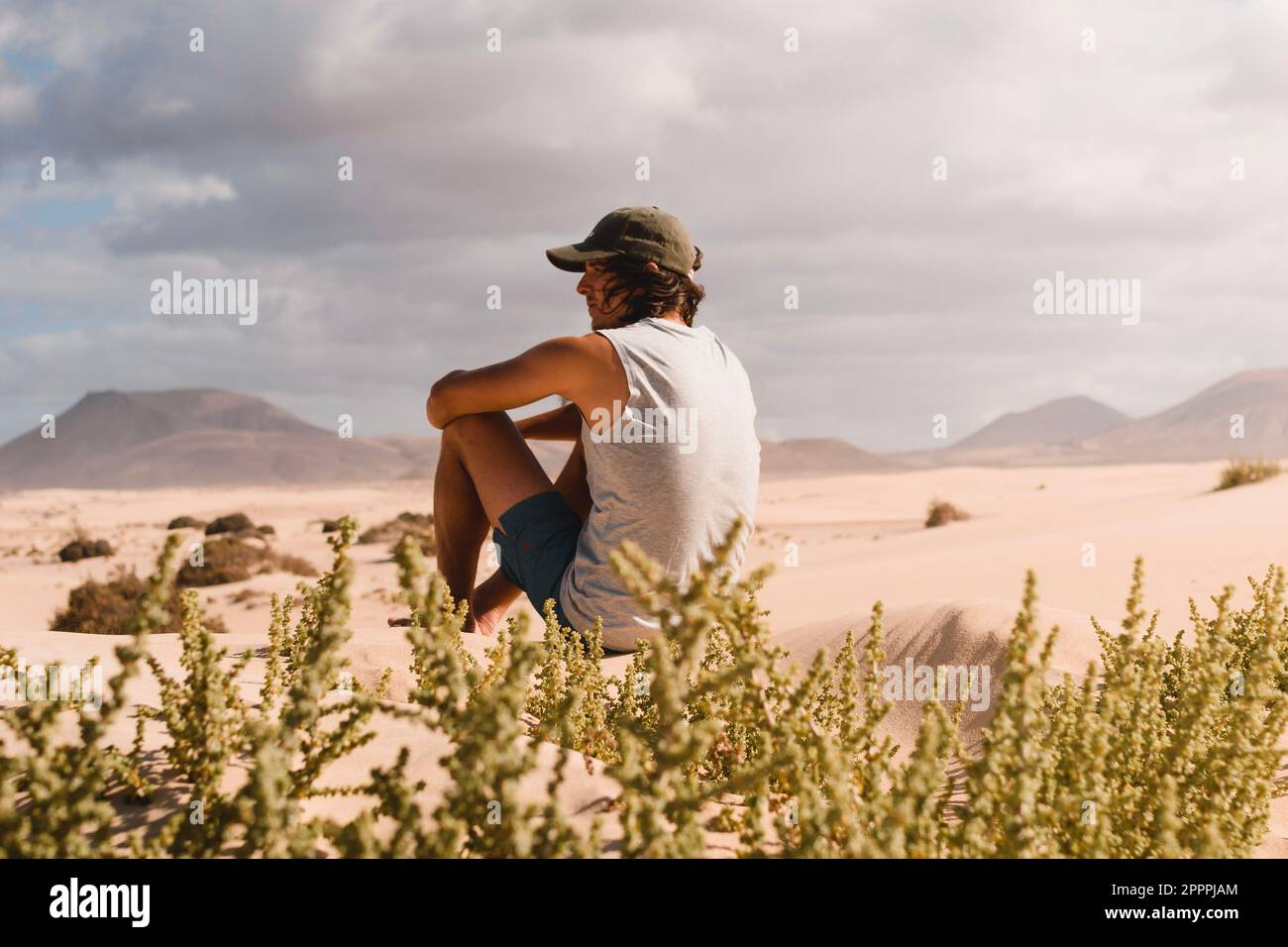 Junger Mann mit Mütze, der auf dem Wüstensand der Dünen von Corralejo in Fuerteventura sitzt und während einer Urlaubsreise den Horizont betrachtet Stockfoto