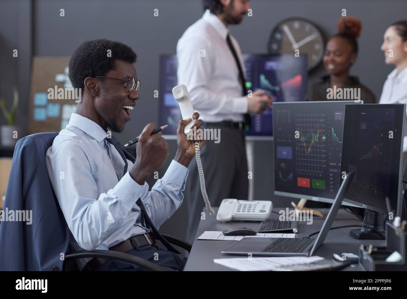 Afroamerikanischer Makler, der mit einem erfolgreichen Geschäft zufrieden war, während er am Telefon mit Computern am Arbeitsplatz saß Stockfoto