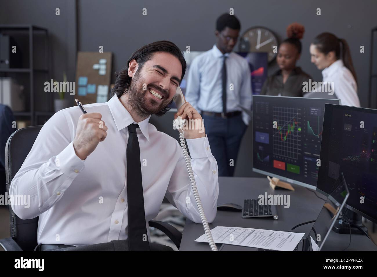 Erfolgreicher Makler, der telefonisch mit ihm spricht und mit dem Geschäft zufrieden ist, während er an seinem Arbeitsplatz mit Computern arbeitet Stockfoto