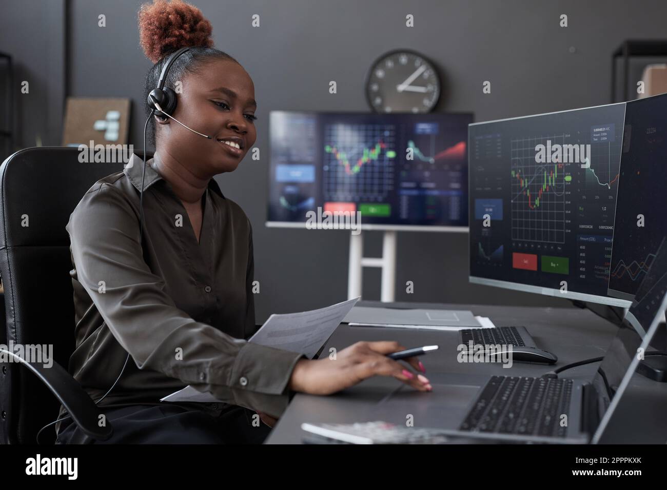 Junge afroamerikanische Maklerin im Kopfhörergeschäft, die mit Kunden spricht, während sie Finanzdiagramme auf Monitoren untersucht, die an ihrem Arbeitsplatz arbeiten Stockfoto