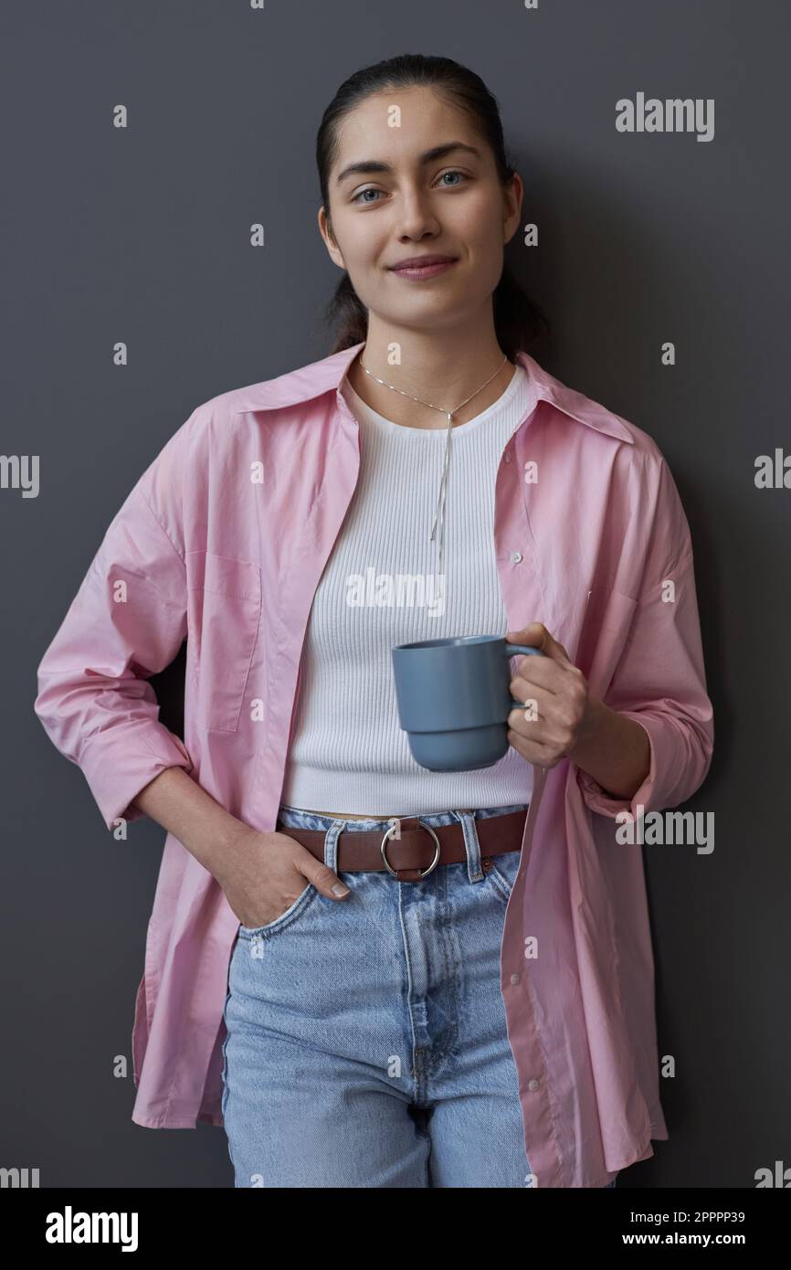 Vertikales Bild einer jungen Frau, die vor der Kamera lächelt und mit einer Tasse Kaffee vor grauem Hintergrund steht Stockfoto