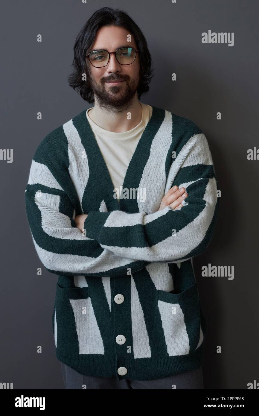 Vertikales Bild eines jungen, gutaussehenden Kerls in Strickjacke, der mit gekreuzten Armen steht und isoliert auf grauen Hintergrund in die Kamera schaut Stockfoto