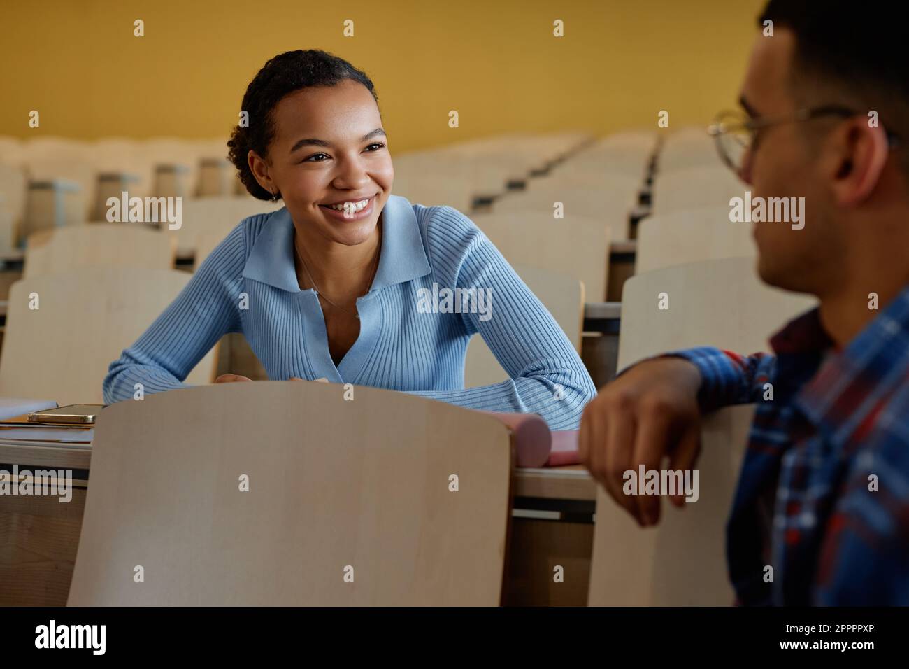 Lächelndes Teenager-Mädchen, das mit ihrer Klassenkameradin spricht, während sie während des Vortrags am Schreibtisch sitzen Stockfoto