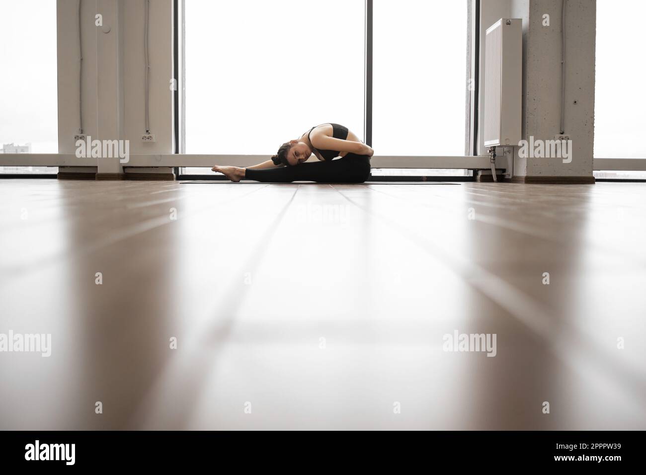 Seitlicher Blick auf die knickige junge Brünette, die sich auf einer Yoga-Matte in der Nähe von Panoramafenstern ausstreckt. Charmante weiße Frau in schwarzer Sportkleidung, die nach dem täglichen Trainingsprogramm fit bleibt. Stockfoto