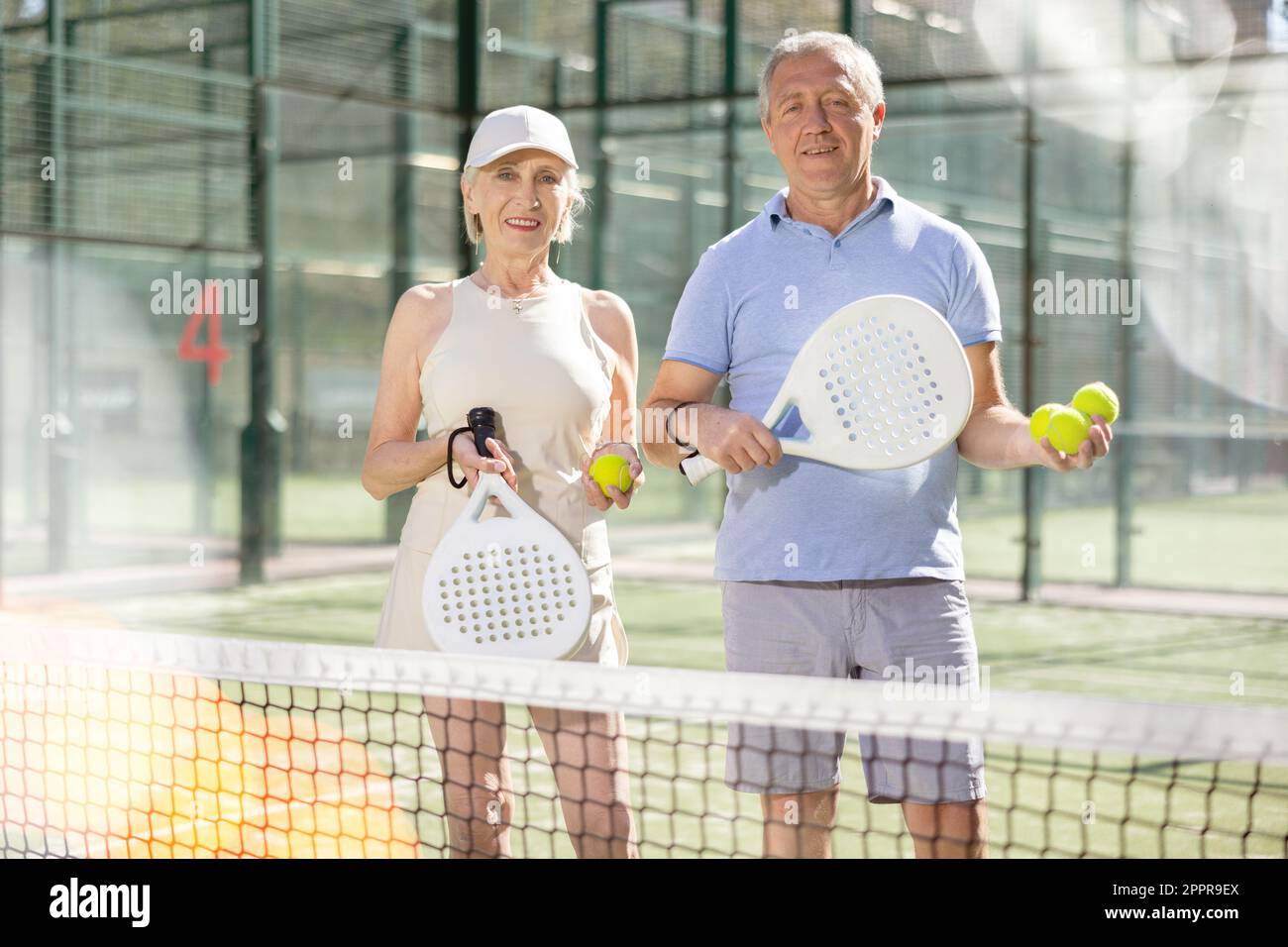 Alter Mann und Frau posieren mit Padel Schläger in den Händen neben dem Netz auf dem Tennisplatz Stockfoto